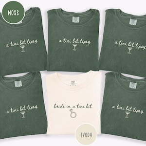 May include: A collection of moss green and ivory t-shirts. The moss green shirts have the text "a tini bit tipsy" with a martini glass graphic. The ivory shirt says "bride in a tini bit" with a ring graphic. The shirts are made of a soft material.