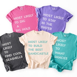 Puede incluir: Cinco camisetas de manga corta y cuello redondo en rosa, morado, gris oscuro, crema y verde azulado. Cada camiseta presenta texto blanco o verde azulado con estilo desgastado con frases como Most Likely To Dig A Big Hole, Stay In The Water, Find Cool Seashells, Build The Best Sandcastle y Want Snacks.