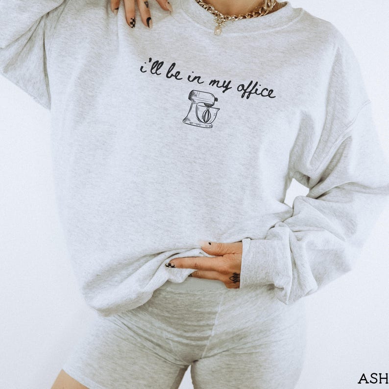 May include: A light gray heather crewneck sweatshirt featuring the black text "i'll be in my office" above a small black line drawing of a stand mixer. The long-sleeved garment has a ribbed collar, cuffs, and hem, providing a casual look.