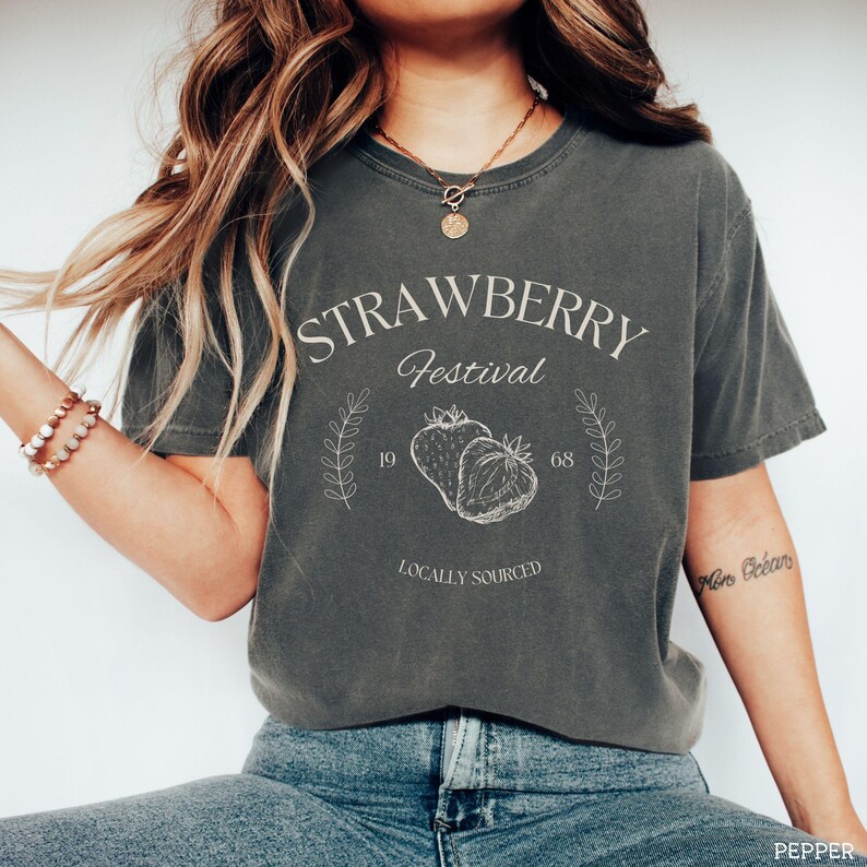 K&ouml;nnte beinhalten: Ein dunkelgraues T-Shirt mit einem wei&szlig;en Grafikdesign, auf dem "Strawberry Festival" steht, sowie eine Zeichnung von zwei Erdbeeren und der Text "Locally Sourced".