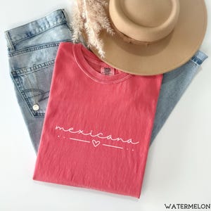 May include: A red t-shirt with the word "mexicana" written in white cursive with a small heart underneath. The shirt is laid out on a pair of blue jeans and a brown hat.