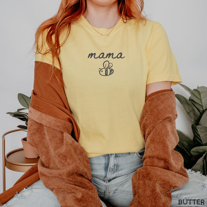 May include: Pale yellow t-shirt featuring the word "mama" in a handwritten style, with a bee graphic. The shirt is made of a soft fabric. The t-shirt is part of a clothing collection.