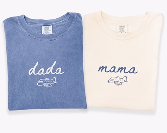 May include: Two short-sleeve t-shirts. The blue jean shirt features the white text 'dada' above a white airplane graphic. The ivory shirt features the blue text 'mama' above a blue airplane graphic. Both shirts have Comfort Colors labels.