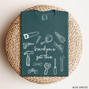 Peut inclure: T-shirt bleu &eacute;pic&eacute;a avec des dessins au trait blancs d'outils, dont une scie, un marteau et une cl&eacute;. Le texte "Grandpa's got this" est au centre. Le t-shirt est pli&eacute; sur une surface circulaire tiss&eacute;e.