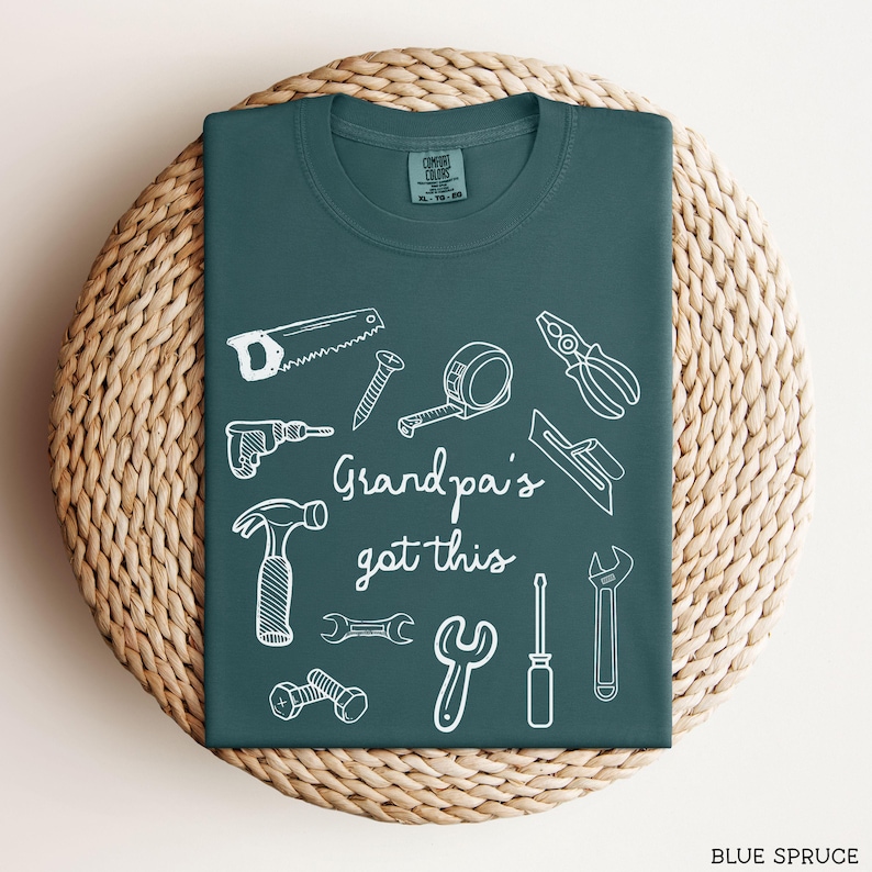 Peut inclure: T-shirt bleu &eacute;pic&eacute;a avec des dessins au trait blancs d'outils, dont une scie, un marteau et une cl&eacute;. Le texte "Grandpa's got this" est au centre. Le t-shirt est pli&eacute; sur une surface circulaire tiss&eacute;e.