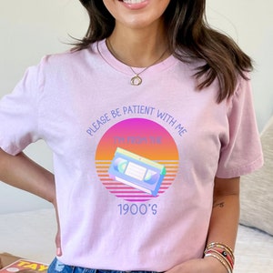 May include: A light pink t-shirt featuring a graphic of a VHS cassette tape centered within a sunset-colored circle. The text reads: PLEASE BE PATIENT WITH ME I'M FROM THE 1900'S. The design uses purple, orange, and yellow hues against the fabric.