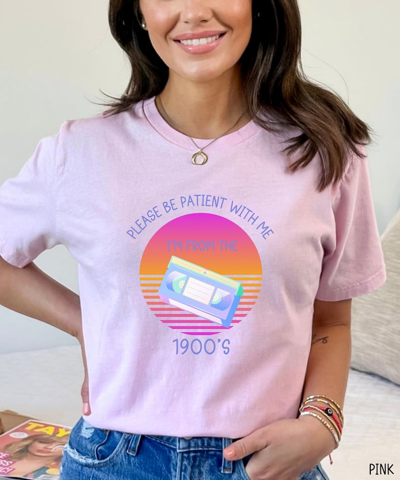 May include: A light pink t-shirt featuring a graphic of a VHS cassette tape centered within a sunset-colored circle. The text reads: PLEASE BE PATIENT WITH ME I'M FROM THE 1900'S. The design uses purple, orange, and yellow hues against the fabric.