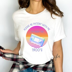 May include: A white t-shirt featuring a graphic print of a VHS cassette tape centered over a circular sunset design with horizontal lines. The text reads, "PLEASE BE PATIENT WITH ME I'M FROM THE 1900'S" in a purple sans-serif font.