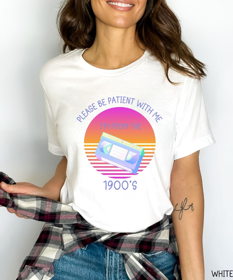 May include: A white t-shirt featuring a graphic print of a VHS cassette tape centered over a circular sunset design with horizontal lines. The text reads, "PLEASE BE PATIENT WITH ME I'M FROM THE 1900'S" in a purple sans-serif font.