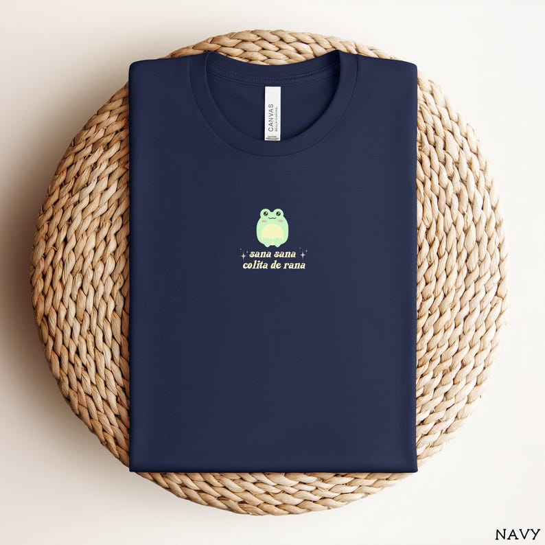 May include: A navy blue t-shirt with a green frog illustration and the text "sana sana colita de rana" in a retro font.