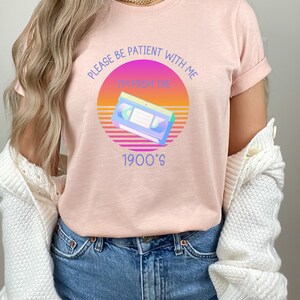 May include: A heather peach t-shirt featuring a graphic of a VHS tape centered over a sunset-colored circle. The text reads, "PLEASE BE PATIENT WITH ME I'M FROM THE 1900'S" in blue lettering. The shirt is paired with blue denim jeans and a white cardigan.