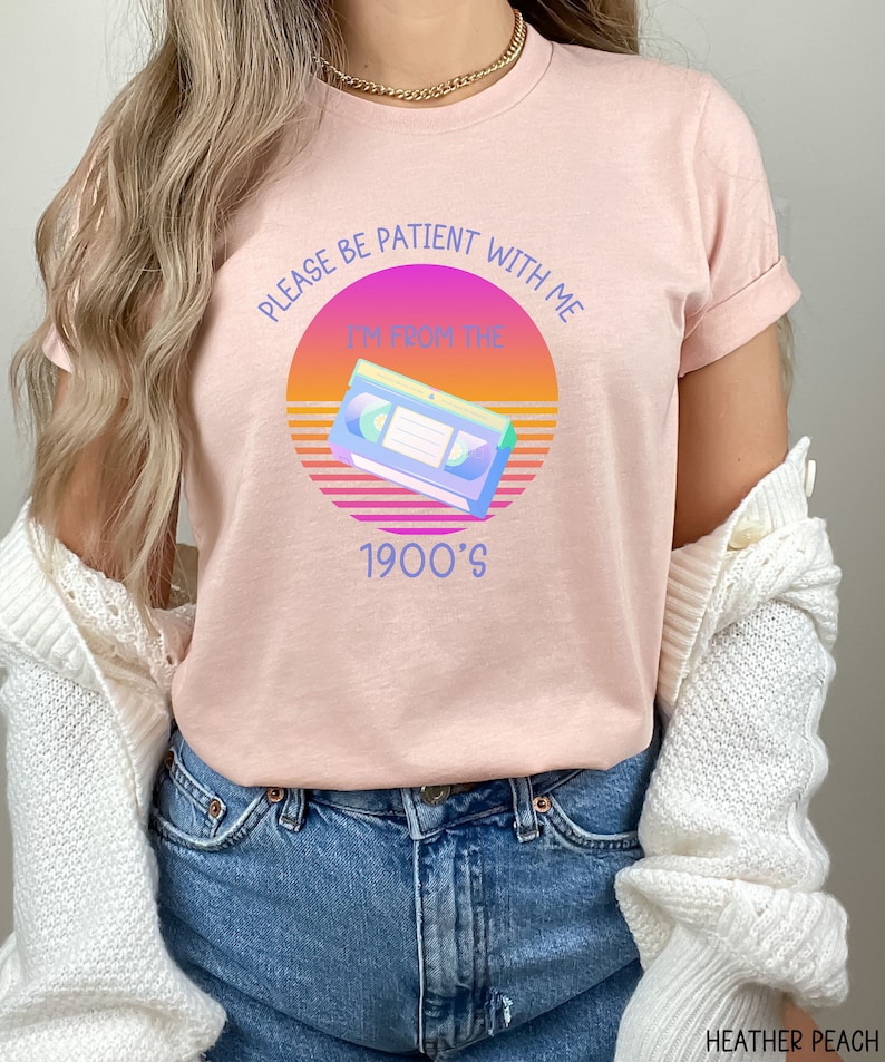 May include: A heather peach t-shirt featuring a graphic of a VHS tape centered over a sunset-colored circle. The text reads, "PLEASE BE PATIENT WITH ME I'M FROM THE 1900'S" in blue lettering. The shirt is paired with blue denim jeans and a white cardigan.