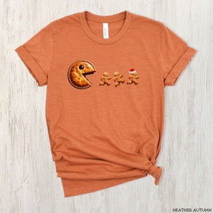 Pu&ograve; includere: Una t-shirt color arancione autunno m&eacute;lange con la grafica di una torta rotonda con un grosso morso, che insegue tre biscotti di pan di zenzero. Uno dei biscotti di pan di zenzero indossa un piccolo cappello da Babbo Natale rosso e bianco.