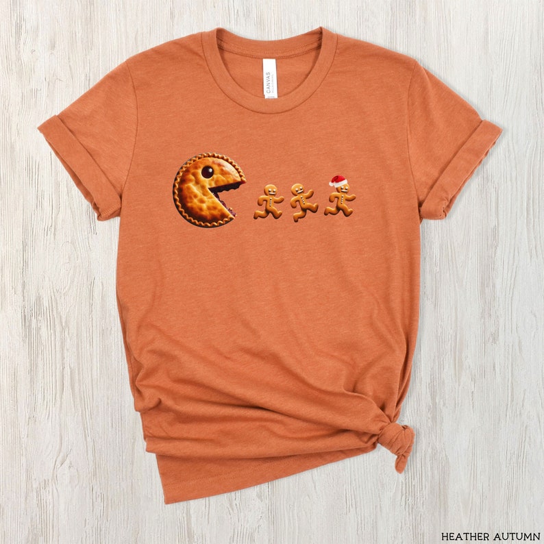 Pu&ograve; includere: Una t-shirt color arancione autunno m&eacute;lange con la grafica di una torta rotonda con un grosso morso, che insegue tre biscotti di pan di zenzero. Uno dei biscotti di pan di zenzero indossa un piccolo cappello da Babbo Natale rosso e bianco.