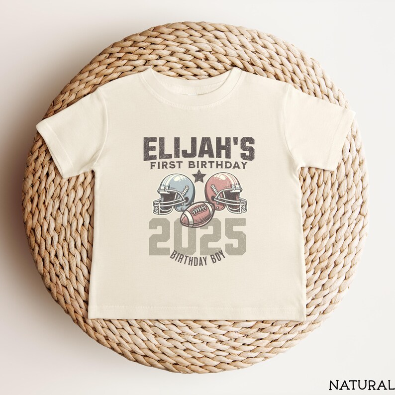 Puede incluir: Una camiseta de manga corta de color natural con un gr&aacute;fico que incluye el texto ELIJAH'S FIRST BIRTHDAY, dos cascos de f&uacute;tbol americano en azul y rojo, un bal&oacute;n de f&uacute;tbol y el a&ntilde;o 2025. El dise&ntilde;o incluye las palabras BIRTHDAY BOY en gris.