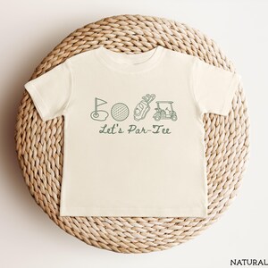 May include: Cream-colored t-shirt featuring a green golf-themed graphic. The design includes a golf flag, ball, golf bag, and golf cart, with the text "Let's Par-Tee" below. The shirt is displayed on a woven, circular surface.