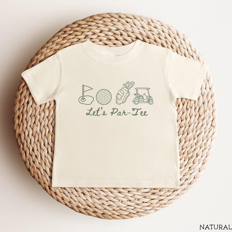 May include: Cream-colored t-shirt featuring a green golf-themed graphic. The design includes a golf flag, ball, golf bag, and golf cart, with the text "Let's Par-Tee" below. The shirt is displayed on a woven, circular surface.