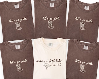 May include: Five brown t-shirts featuring the white embroidered text let's go girls and a cowboy boot graphic, and one white t-shirt featuring the black embroidered text man, i feel like i'm 40 and a cowboy hat graphic, all with crew necklines.
