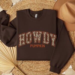 May include: A dark brown crewneck sweatshirt featuring the text HOWDY in large block letters with a tan and brown western-themed pattern inside the characters, and the word PUMPKIN printed in orange underneath the main text.