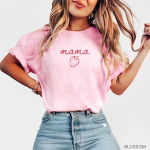 May include: A light pink short-sleeved t-shirt featuring the word mama in a dark pink cursive font centered above a small dark pink strawberry graphic. The shirt is paired with blue denim jeans that have a visible button fly and metal rivets.
