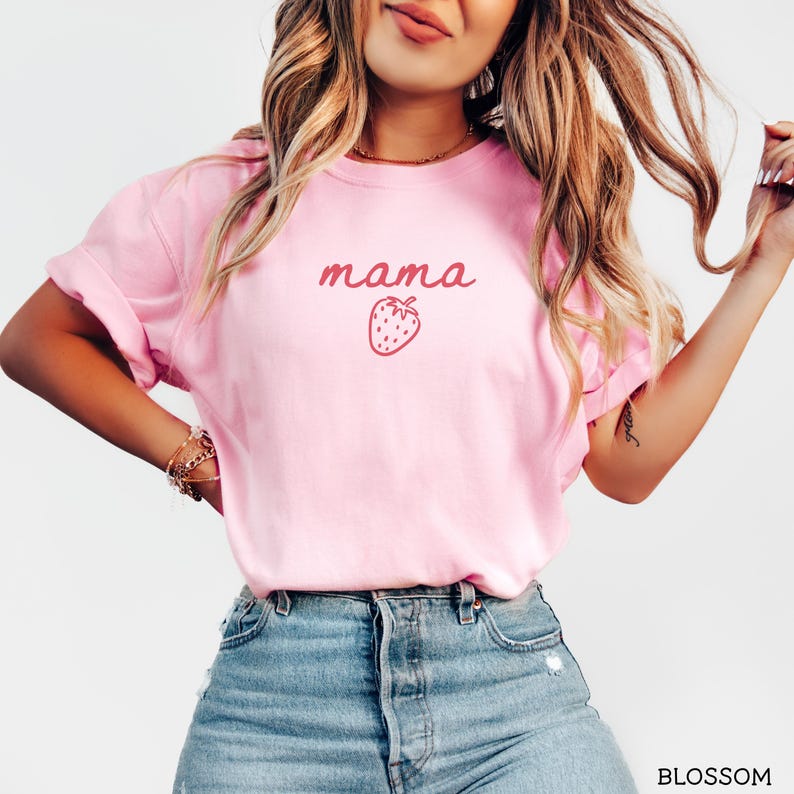 May include: A light pink short-sleeved t-shirt featuring the word mama in a dark pink cursive font centered above a small dark pink strawberry graphic. The shirt is paired with blue denim jeans that have a visible button fly and metal rivets.