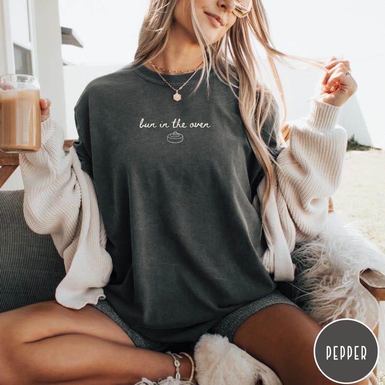 K&ouml;nnte beinhalten: Dunkelgraues T-Shirt mit dem Schriftzug "bun in the oven" in wei&szlig;er Schrift und einer kleinen Grafik eines Br&ouml;tchens. Das Shirt hat eine lockere Passform und scheint aus einem weichen Material zu sein. Teil einer Schwangerschaftsank&uuml;ndigungs-Kollektion.