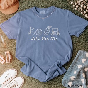 May include: A blue jean colored t-shirt with a white graphic of golf-related images and the text "Let's Par-Tee". The shirt is surrounded by a brown hat, white sneakers, and a blue towel with white hearts.