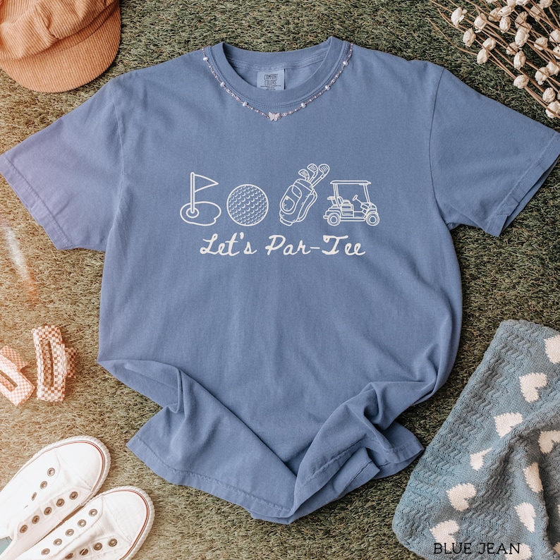 May include: A blue jean colored t-shirt with a white graphic of golf-related images and the text "Let's Par-Tee". The shirt is surrounded by a brown hat, white sneakers, and a blue towel with white hearts.