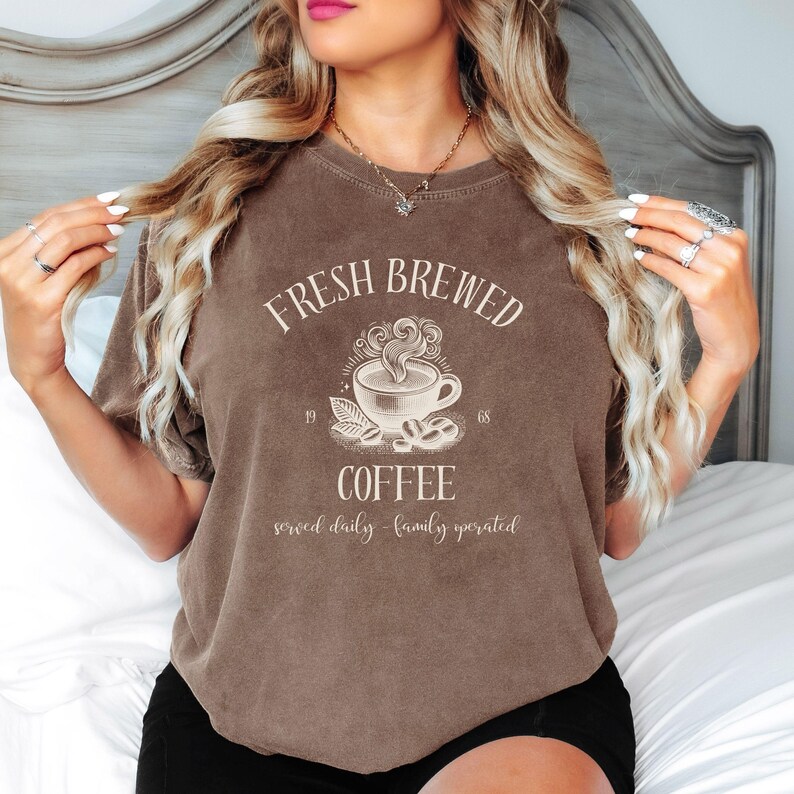 Op de afbeelding: Een bruin T-shirt met korte mouwen en een cr&egrave;mekleurige afbeelding van een dampende kop koffie, de tekst "FRESH BREWED COFFEE" en "served daily - family operated", en de cijfers 19 en 68 aan weerszijden van de koffiebonen.