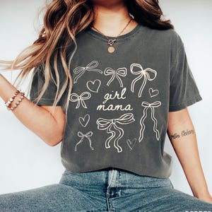 May include: A dark gray t-shirt with a graphic design of bows, hearts, and the text "girl mama".
