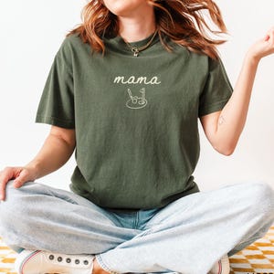 May include: A woman wearing a dark green t-shirt with the text "mama" and a golf club and ball graphic. She is wearing light blue jeans and white sneakers.