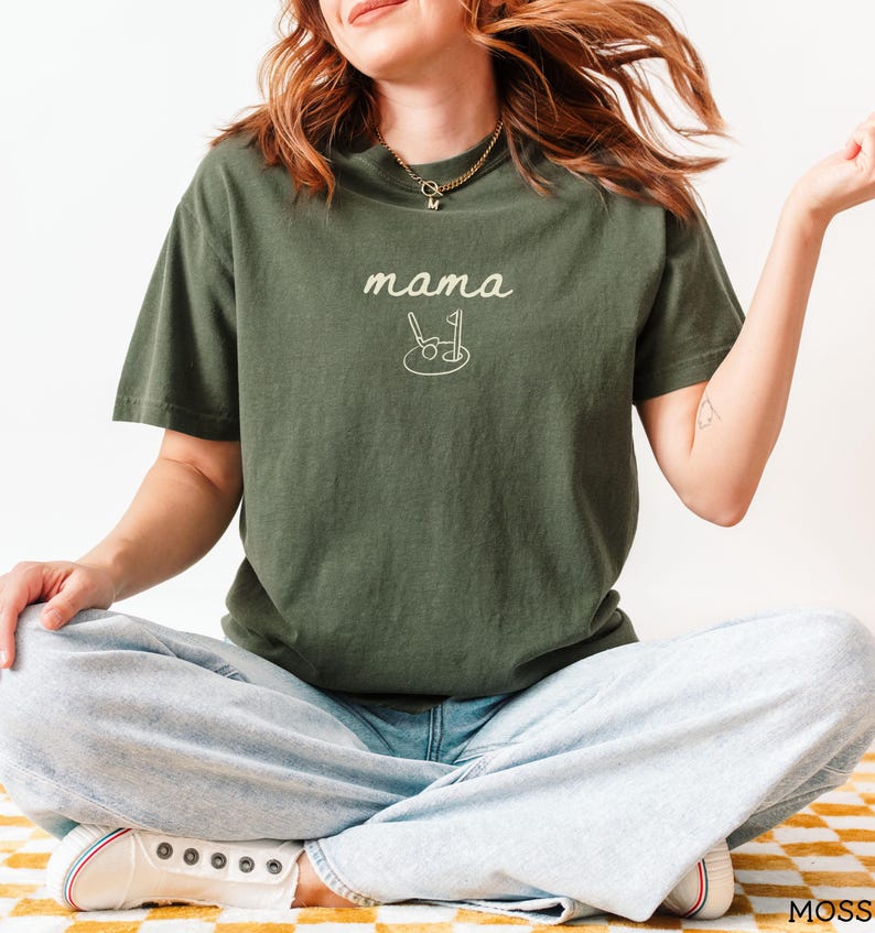 May include: A woman wearing a dark green t-shirt with the text "mama" and a golf club and ball graphic. She is wearing light blue jeans and white sneakers.