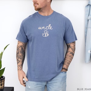 May include: A blue t-shirt featuring the word "uncle" in a cursive font, with a bee graphic beneath it. The shirt is a solid blue colour and has short sleeves. The text and bee are in a light beige colour.