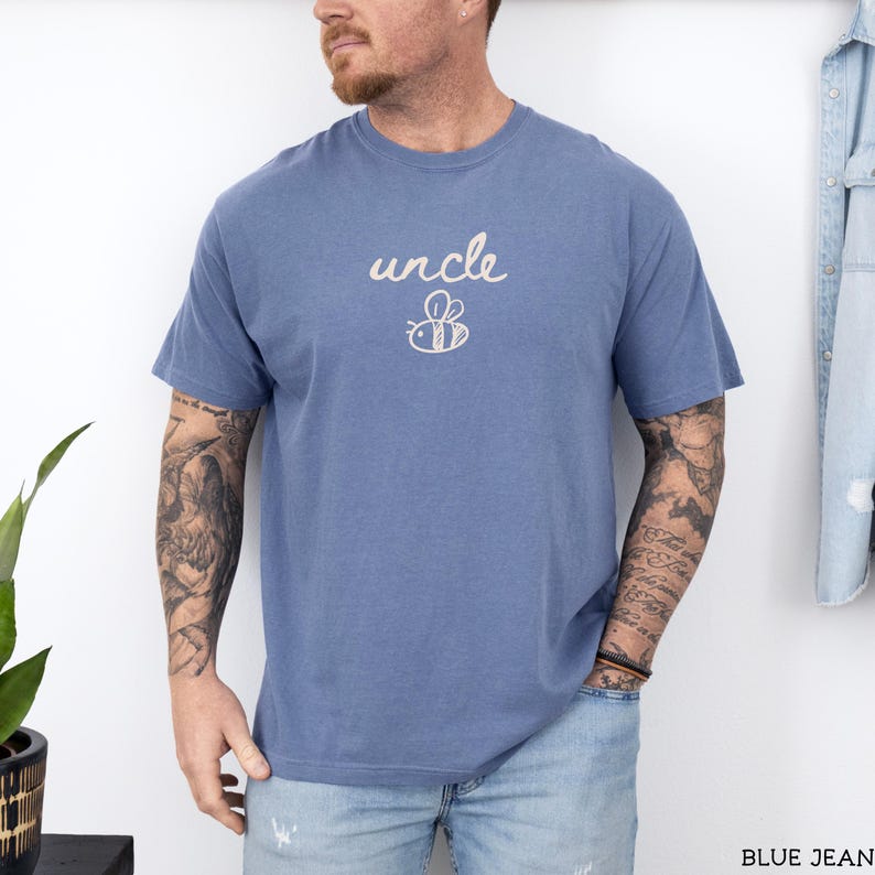 May include: A blue t-shirt featuring the word "uncle" in a cursive font, with a bee graphic beneath it. The shirt is a solid blue colour and has short sleeves. The text and bee are in a light beige colour.