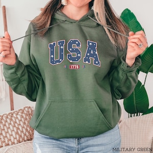 K&ouml;nnte beinhalten: Milit&auml;rgr&uuml;ner Kapuzenpullover mit Fronttasche und Kordelzug. Das Wort "USA" ist in einem blau-wei&szlig;en Sternmuster auf die Brust gedruckt, darunter "1776". Der Hoodie ist aus weichem Material.