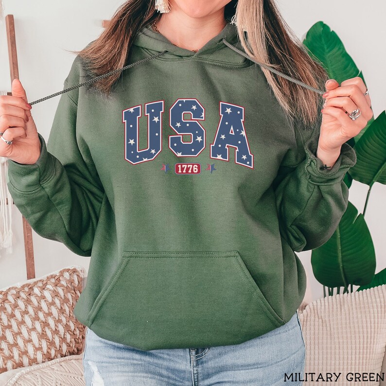 K&ouml;nnte beinhalten: Milit&auml;rgr&uuml;ner Kapuzenpullover mit Fronttasche und Kordelzug. Das Wort "USA" ist in einem blau-wei&szlig;en Sternmuster auf die Brust gedruckt, darunter "1776". Der Hoodie ist aus weichem Material.