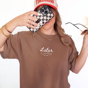 Pu&ograve; includere: T-shirt marrone con la scritta "Later" e una grafica di coccodrillo. Cappellino da baseball beige e rosso con la scritta "TIMELESS". Custodia per telefono a scacchi bianchi e neri.