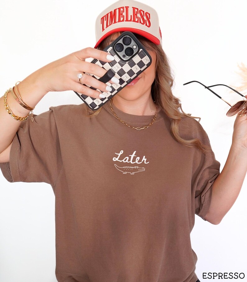 Pu&ograve; includere: T-shirt marrone con la scritta "Later" e una grafica di coccodrillo. Cappellino da baseball beige e rosso con la scritta "TIMELESS". Custodia per telefono a scacchi bianchi e neri.