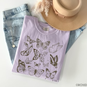 May include: A lavender t-shirt with a graphic print of butterflies in shades of brown. The text "COMFORT COLORS" is printed on the shirt.