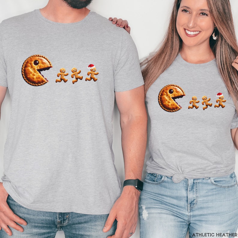 Pu&ograve; includere: Una t-shirt grigio chiaro con una grafica raffigurante una grande torta rotonda color marrone dorato con un occhio solo, che sembra inseguire tre piccoli omini di pan di zenzero. Uno degli omini indossa un piccolo cappello da Babbo Natale.