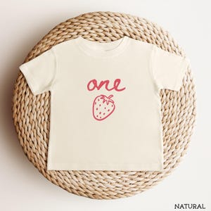 May include: A natural-colored short-sleeve t-shirt featuring the word one written in a pink cursive script above a simple pink line-art illustration of a strawberry centered on the front of the garment.