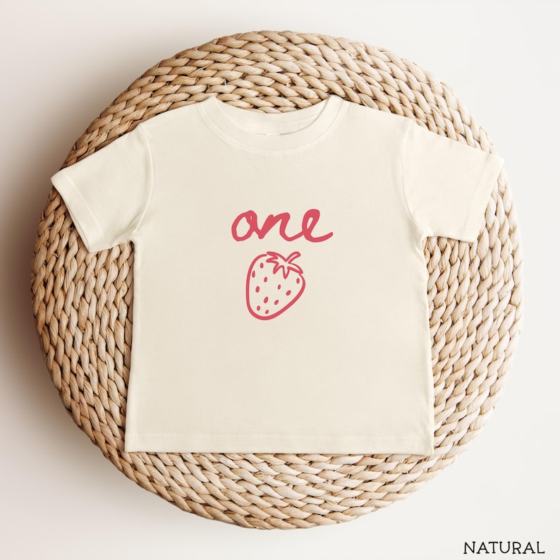 May include: A natural-colored short-sleeve t-shirt featuring the word one written in a pink cursive script above a simple pink line-art illustration of a strawberry centered on the front of the garment.