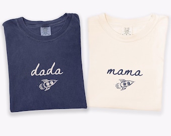 Puede incluir: Dos camisetas, una azul marino y otra color marfil. La camiseta azul marino presenta el texto blanco "dada" sobre un gráfico de cohete blanco. La camiseta color marfil presenta el texto azul marino "mama" sobre un gráfico de cohete.