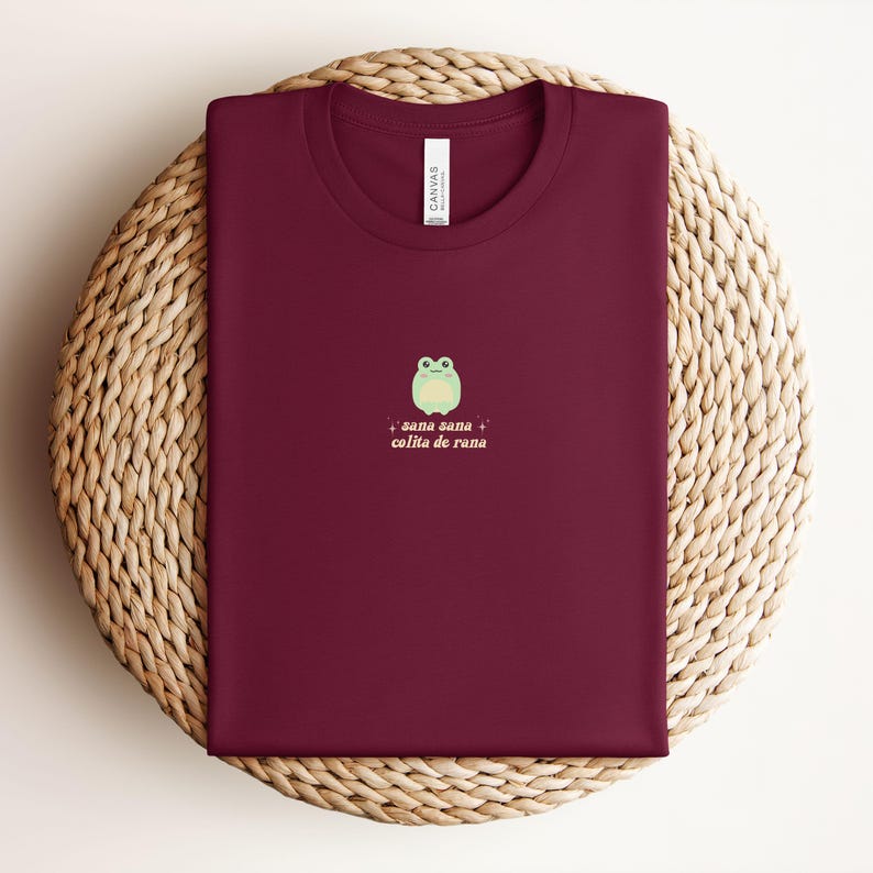 May include: A maroon t-shirt with a green frog graphic and the text "sana sana colita de rana" in pink and white.