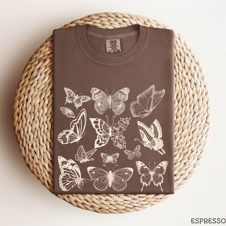 May include: A brown t-shirt with a white print of various butterflies. The butterflies are in different sizes and positions, creating a whimsical and organic design.