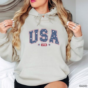K&ouml;nnte beinhalten: Ein sandfarbener Kapuzenpullover mit Fronttasche und Kordelzugkapuze. Das Wort "USA" ist in blauer Schrift mit wei&szlig;en Sternen, rot umrandet, auf die Brust gedruckt. Unter dem Wort "USA" steht die Zahl "1776" in einem roten Rechteck.