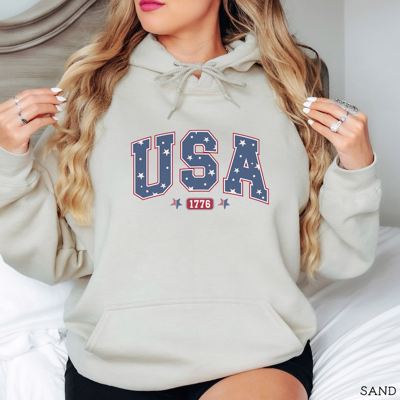 K&ouml;nnte beinhalten: Ein sandfarbener Kapuzenpullover mit Fronttasche und Kordelzugkapuze. Das Wort "USA" ist in blauer Schrift mit wei&szlig;en Sternen, rot umrandet, auf die Brust gedruckt. Unter dem Wort "USA" steht die Zahl "1776" in einem roten Rechteck.