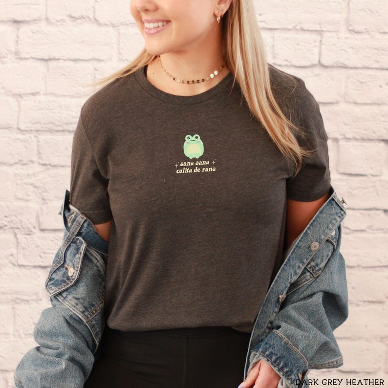 May include: A dark grey heather t-shirt with a green frog illustration and the text "sana sana colita de rana".
