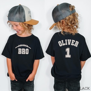May include: A black t-shirt featuring a white football graphic with the text BRO on the front and the name OLIVER with the number 1 on the back, paired with a grey and tan trucker hat with a mesh back and a solid tan brim.
