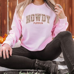 May include: A light pink long-sleeve sweatshirt featuring the text HOWDY PUMPKIN printed on the front. The word HOWDY is displayed in a large, block-style font with a tan and brown patterned fill, while the word PUMPKIN appears below in solid brown.